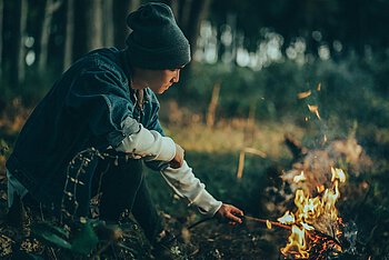 Junger Mann hält Stock in ein Lagerfeuer. Er befindet sich im Wald.